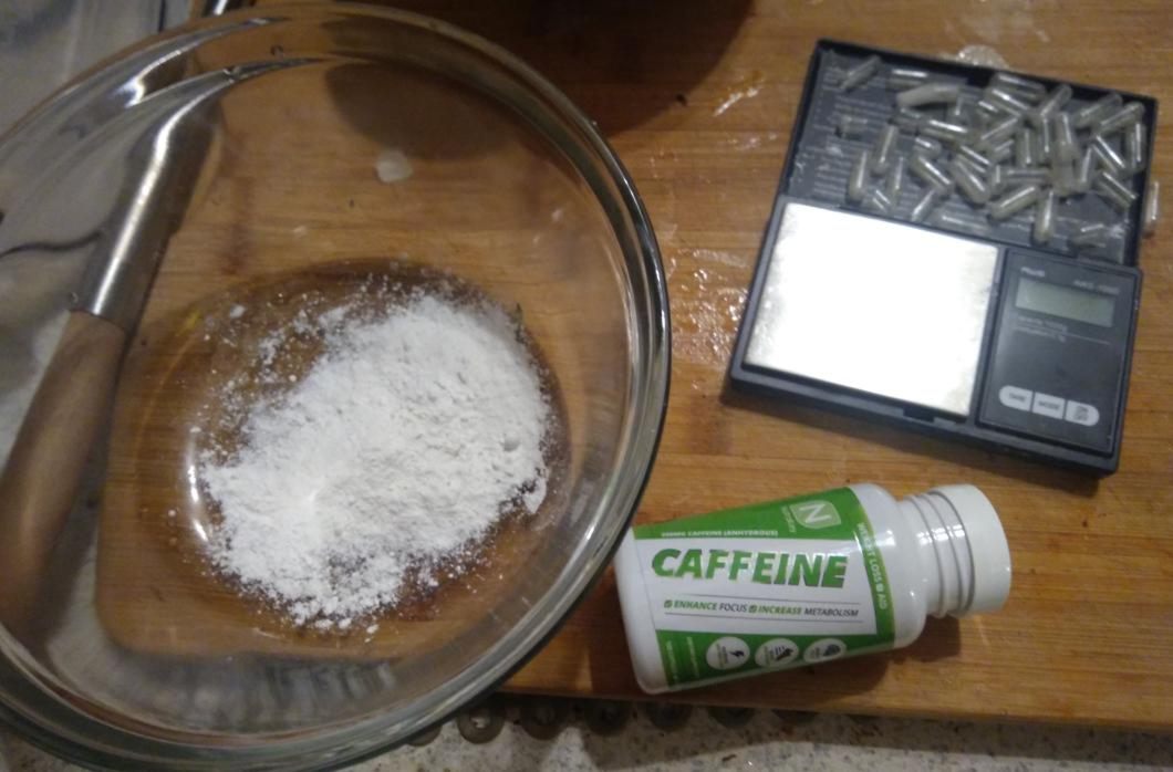 a pile of empty gel caps sitting next to a digital scale, a bottle of caffeine pills, and a little mound of white powder in a glass bowl
