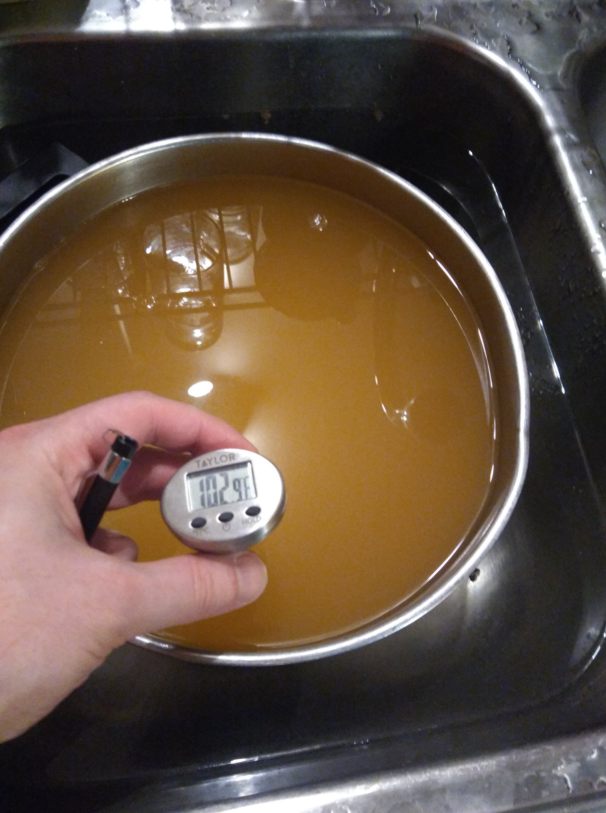 a pot of tan or brown liquid sits in a sink basin filled with water. The photographer is measuring the temperature of the liquid at 102 degrees Fahrenheit 