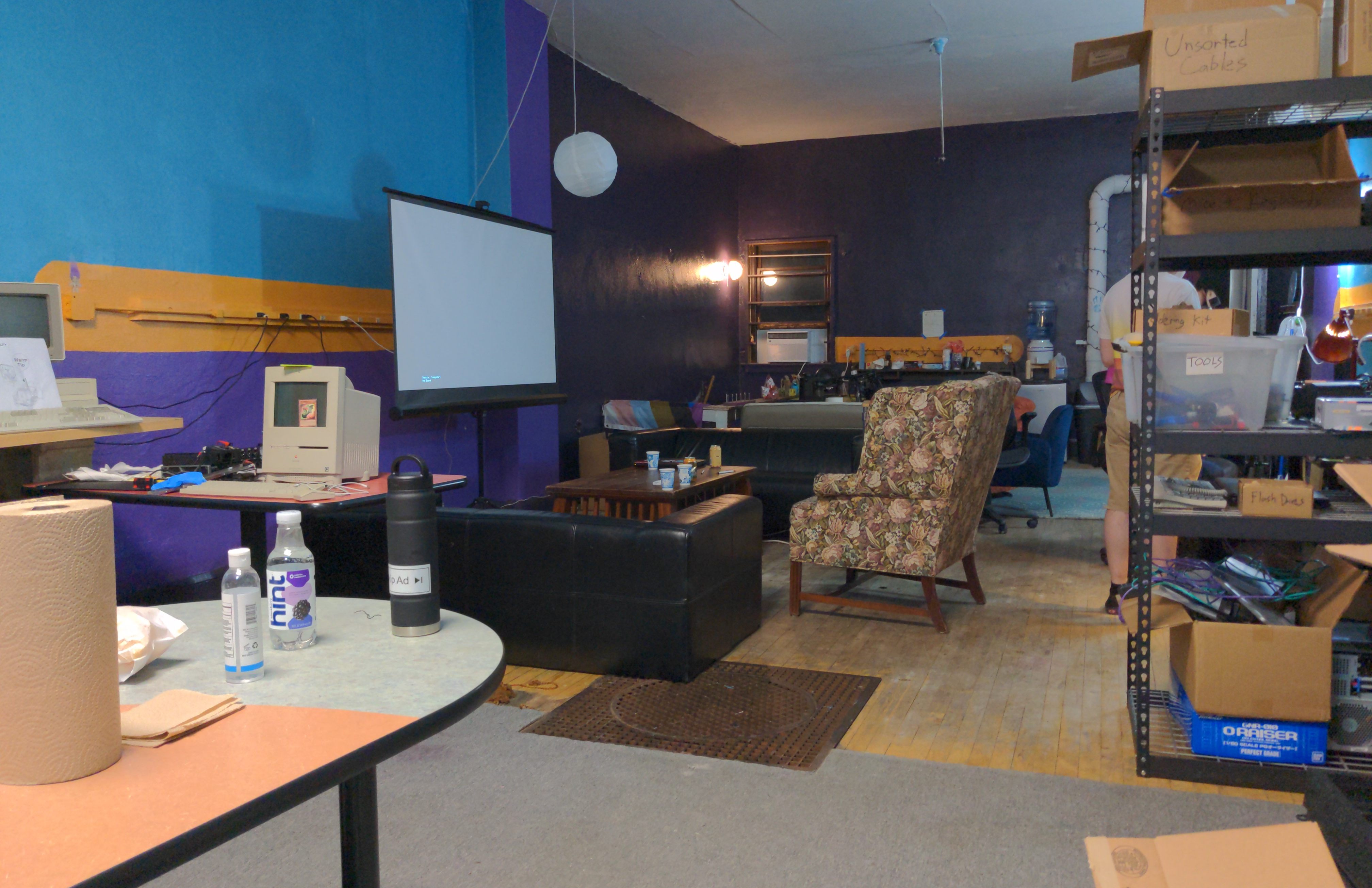 Photograph of the Layer Zero hackerspace featuring comfy padded armchairs and sofas,  a steel shelving unit, a projector screen, and workbenches with chairs.