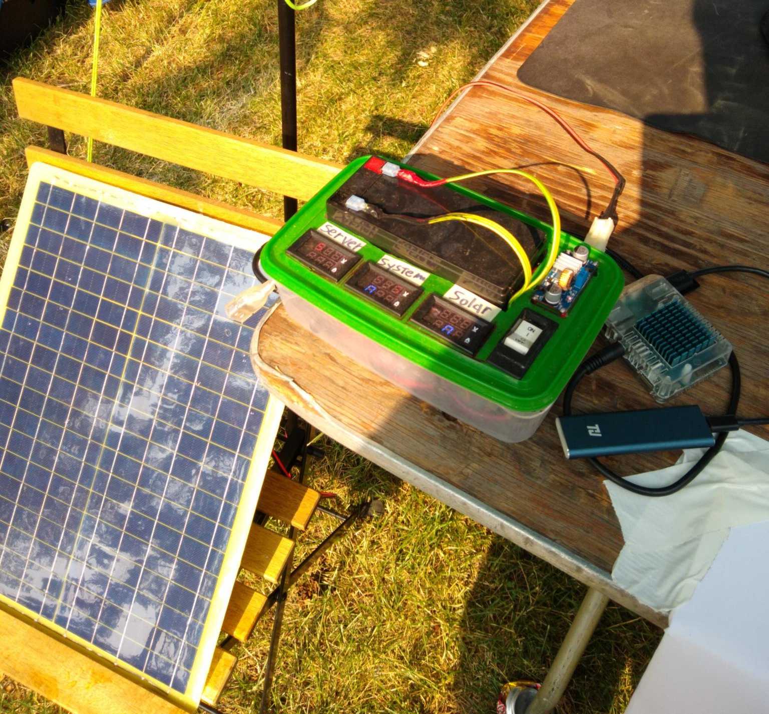 Sparse whispy clouds are visible reflected off of a dark blue and green photovoltaic panel resting on a wood and steel IKEA lawn chair.  A lead acid battery, a large safety switch, and a small Buck Converter are sticking up through  holes cut in a green tupperware container.  There are three voltage/amperage displays labeled "Server",  "System", and "Solar"."