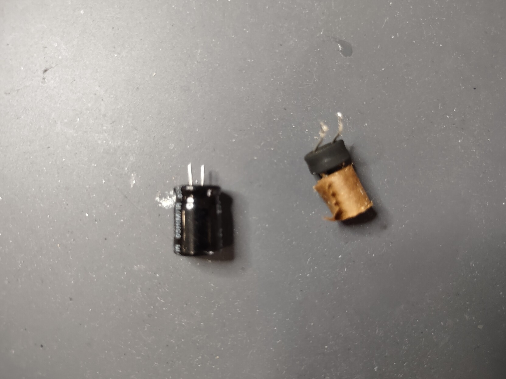 Coseup photo of the two capacitors on the solder mat at Layer Zero. The exploded one looks like a brown roll of paper with a rubber bung on the bottom and two long bent wires sticking out.  The replacement is still in its shiny black metal casing with to very short straight wires.